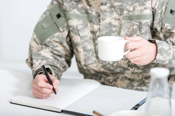 Abgeschnittene Ansicht eines Mannes in Militäruniform, der Kaffee trinkt und in Notizbuch schreibt — Stockfoto