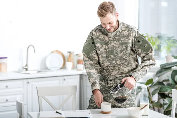 Hombre en uniforme de camuflaje verter café en taza en la cocina - foto de stock