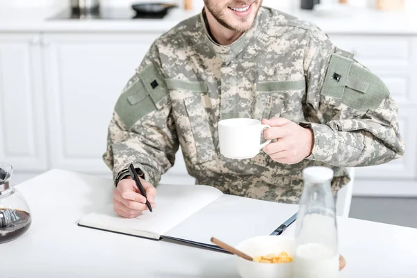 Homem de uniforme militar bebe café e escreve em caderno — Fotografia de Stock