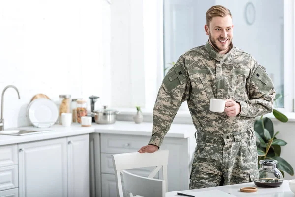 Soldado do exército sorrindo segurando xícara de café na cozinha — Fotografia de Stock