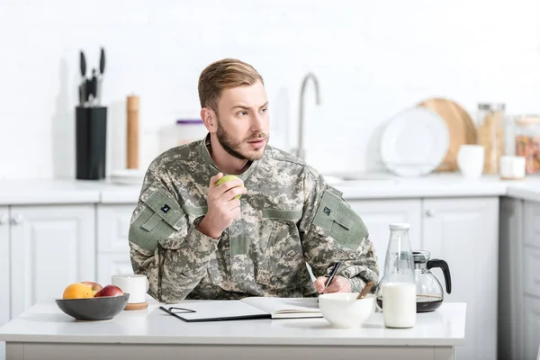 Army soldier sitting at kitchen table and eating apple — Stock Photo