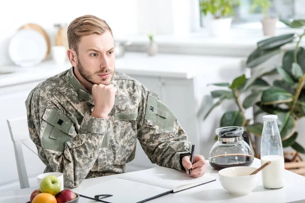 Pensive army soldier sitting at kitchen table and writing while having breakfast — Stock Photo