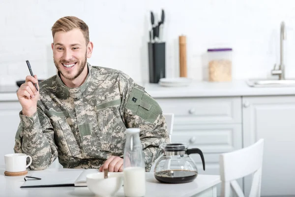 Улыбающийся солдат, сидящий за кухонным столом с ручкой и блокнотом за завтраком — стоковое фото