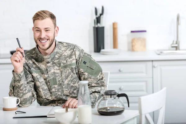 Soldado do exército sorridente sentado na mesa da cozinha com caneta e caderno enquanto toma café da manhã — Fotografia de Stock
