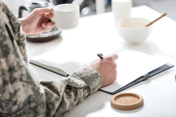 Vista recortada del soldado del ejército sentado en la mesa de la cocina, escribiendo y desayunando - foto de stock