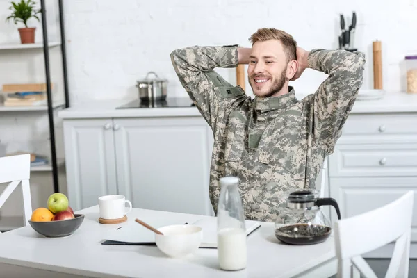 Soldat souriant de l'armée assis à la table de cuisine avec les mains sur la tête tout en prenant le petit déjeuner — Photo de stock