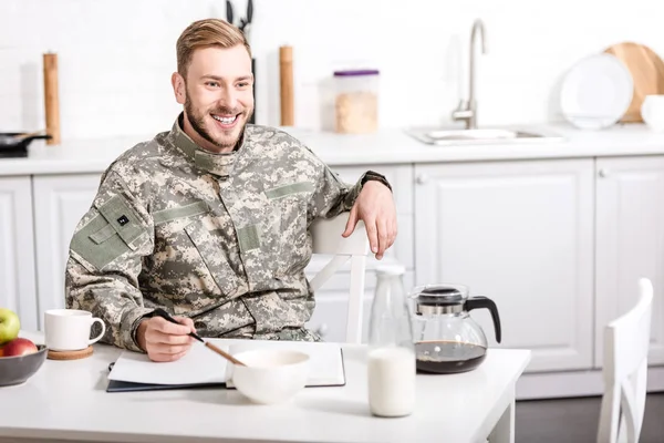 Soldado del ejército sonriente sentado en la mesa de la cocina y desayunando - foto de stock