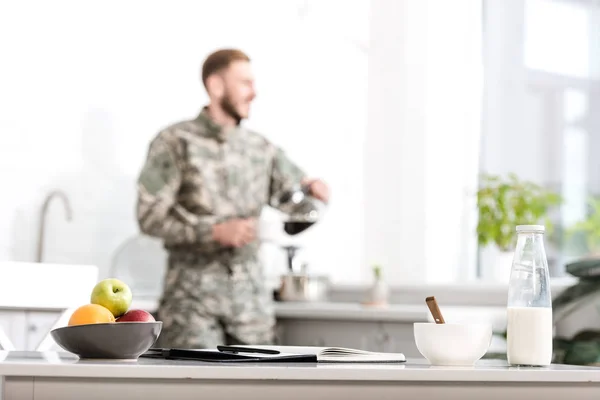 Concentration sélective de la table avec bol de fruits, bouteille de lait, cahier et stylo, soldat de l'armée versant du café filtré dans la cuisine sur le fond — Photo de stock