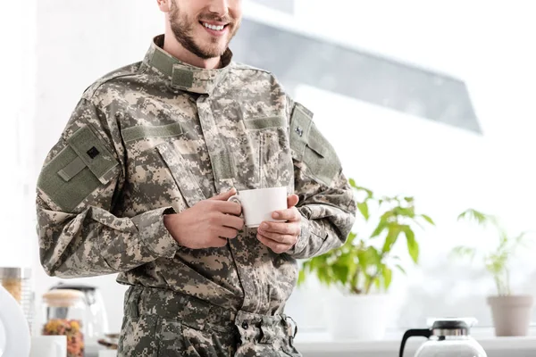 Vista recortada de soldado del ejército sosteniendo taza de café en la cocina - foto de stock