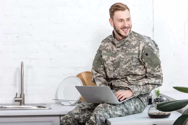 Soldado sonriente en uniforme militar sentado en la encimera de la cocina y utilizando el ordenador portátil - foto de stock