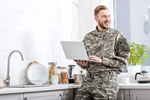 Soldat souriant de l'armée utilisant un ordinateur portable à la cuisine et regardant loin — Photo de stock