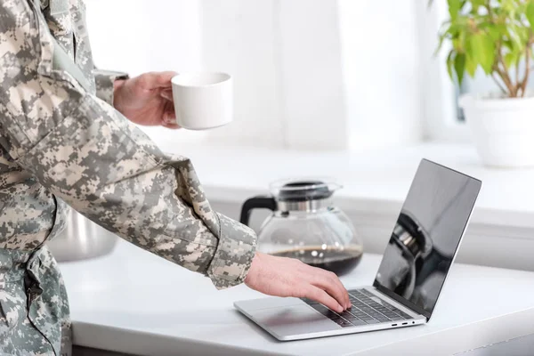 Vista parcial del soldado sosteniendo la taza de café y utilizando el ordenador portátil en la cocina - foto de stock