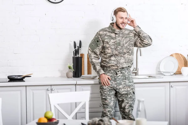 Soldato sorridente in uniforme militare indossando cuffie e ascoltando musica in cucina — Foto stock