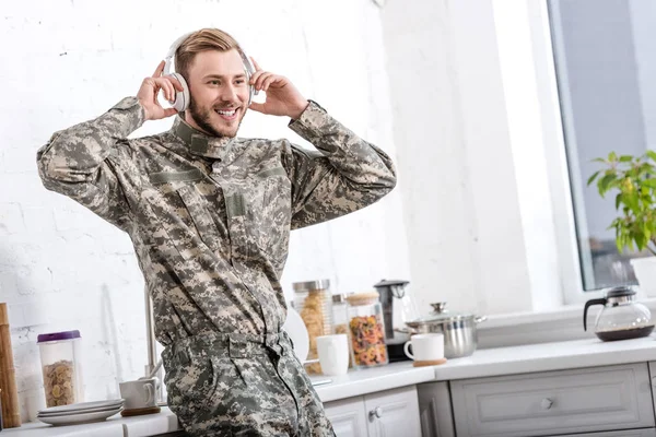 Handsome soldier in military uniform wearing headphones and listening music at kitchen — Stock Photo