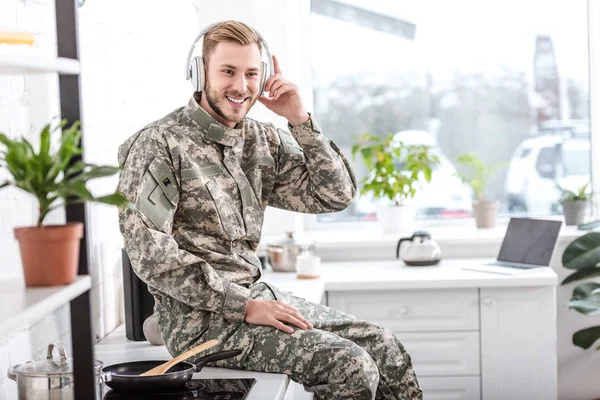 Guapo soldado en auriculares escuchando música y sentado en la encimera de la cocina en casa - foto de stock