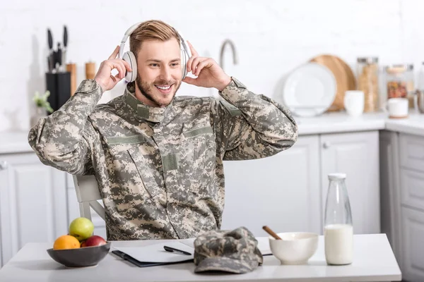 Красивый солдат армии в наушниках сидит за кухонным столом во время завтрака дома — стоковое фото