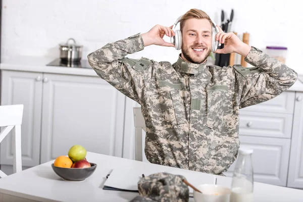 Soldado do exército sorridente em fones de ouvido sentado na mesa da cozinha durante o café da manhã em casa — Fotografia de Stock