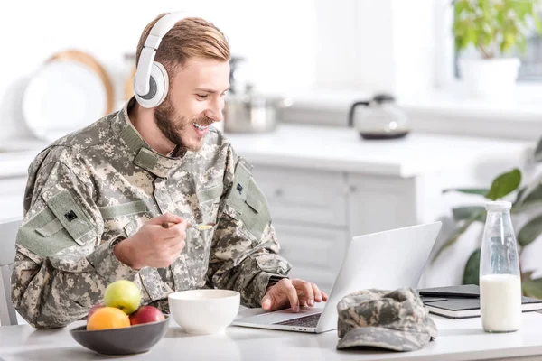 Handsome soldier in headphones using laptop at kitchen table while having breakfast — Stock Photo