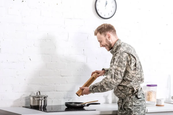 Soldado do exército sorrindo usando panela de pimenta enquanto cozinha na cozinha — Fotografia de Stock