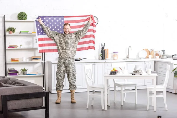 Lächelnder Armeesoldat, der in die Kamera blickt und stolz die amerikanische Flagge in der Küche hält — Stockfoto