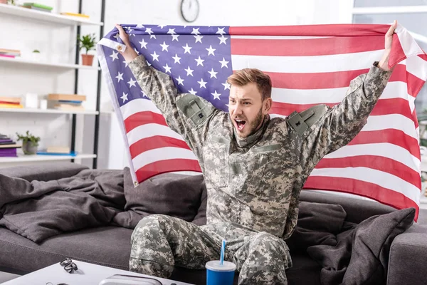 Soldato dell'esercito eccitato seduto sul divano, applaudendo e tenendo orgogliosamente bandiera americana — Foto stock