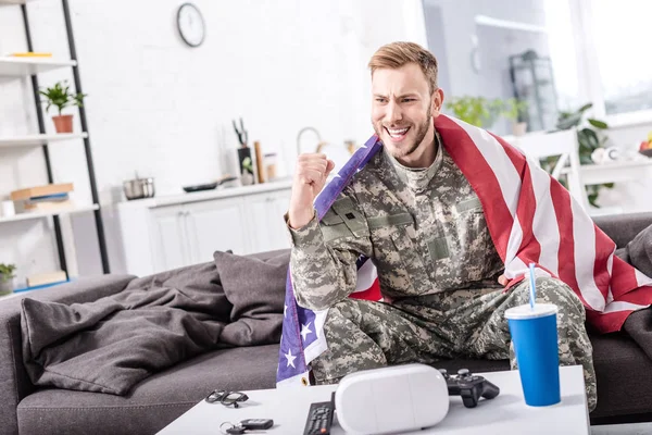 Excited army soldier covered in american flag sitting on couch, cheering and and watching football match at home — Stock Photo