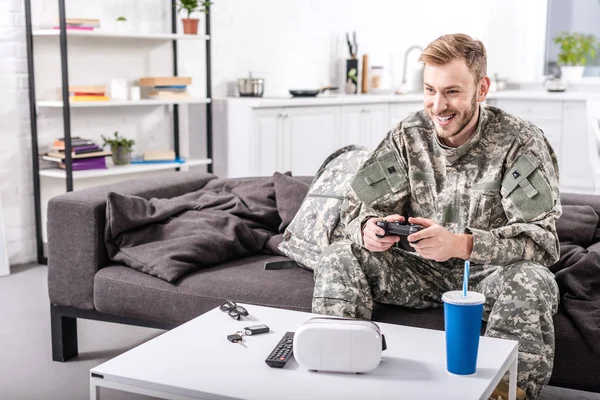 Smiling army soldier sitting on couch at home and playing video game — Stock Photo
