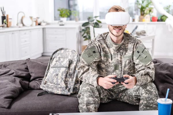 Улыбающийся солдат армии в наушниках виртуальной реальности, играющий в видеоигры на диване — стоковое фото