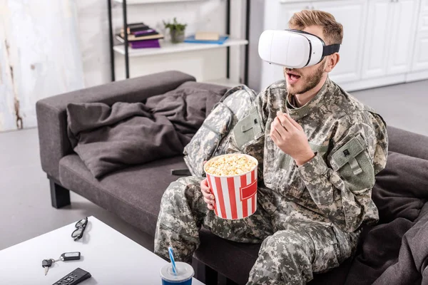 Солдат в наушниках виртуальной реальности, сидит на диване, смотрит кино и ест попкорн — стоковое фото