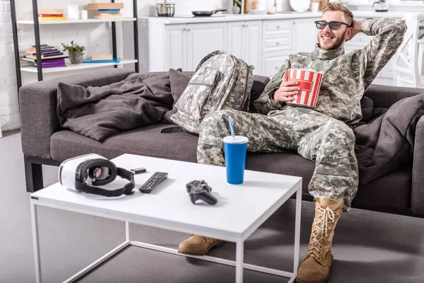 Army soldier in military uniform wearing 3d glasses, eating popcorn and watching movie on couch — Stock Photo