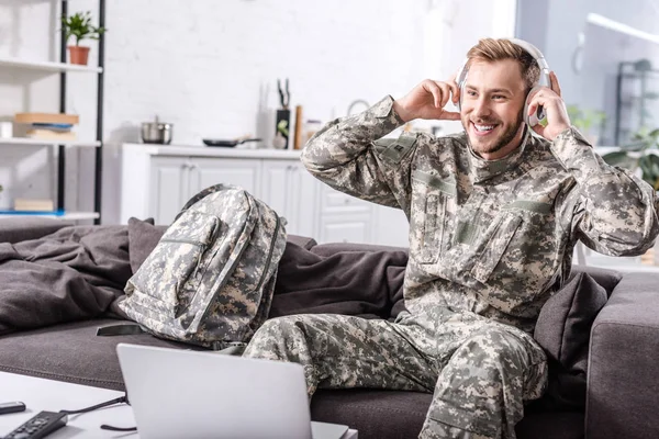 Smiling army soldier in headphones sitting on couch and using laptop at home — Stock Photo