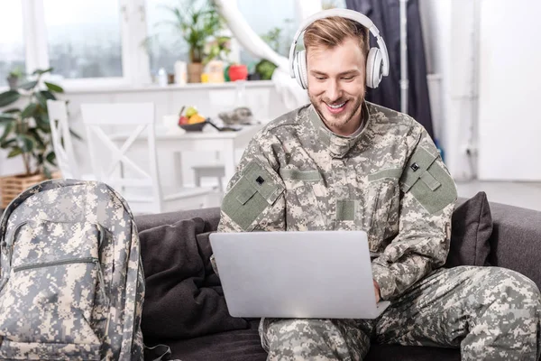 Soldato dell'esercito sorridente indossa le cuffie e utilizzando il computer portatile sul divano — Foto stock