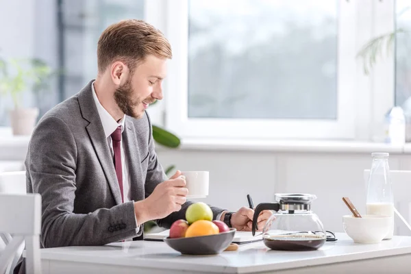 Bonito empresário beber café na cozinha e escrever no bloco de notas — Fotografia de Stock