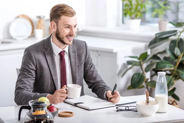Smiling businessman writing in notebook while drinking coffee and having breakfast at home — Stock Photo