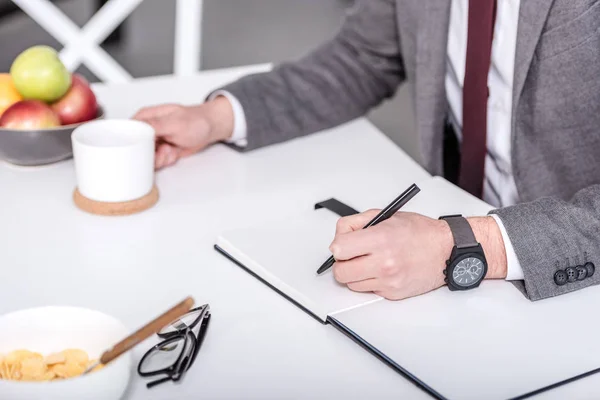 Visão cortada de empresário escrevendo em caderno com caneta enquanto toma café da manhã — Fotografia de Stock