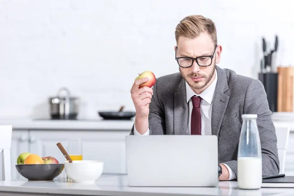 Businessman working on laptop while eating apple at kitchen — Stock Photo