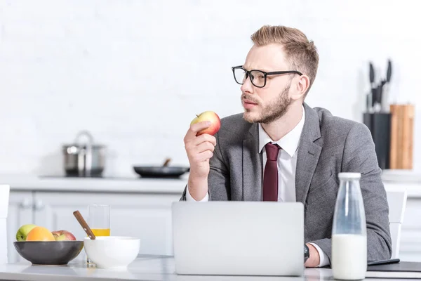 Бизнесмен работает на ноутбуке во время еды яблоко на кухне — стоковое фото