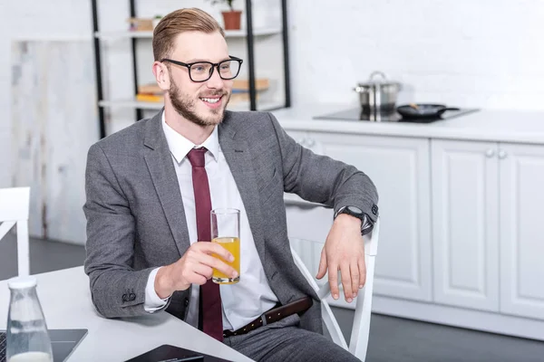 Homem de negócios de óculos sentado na mesa da cozinha, bebendo suco de laranja e olhando para longe — Fotografia de Stock