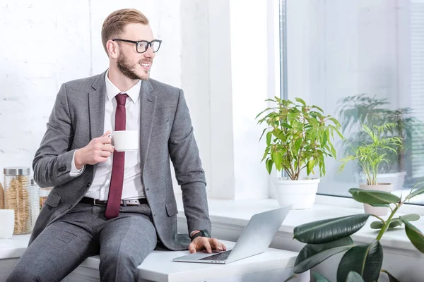 Businessman in glasses sitting on countertop and drinking coffee in kitchen — Stock Photo