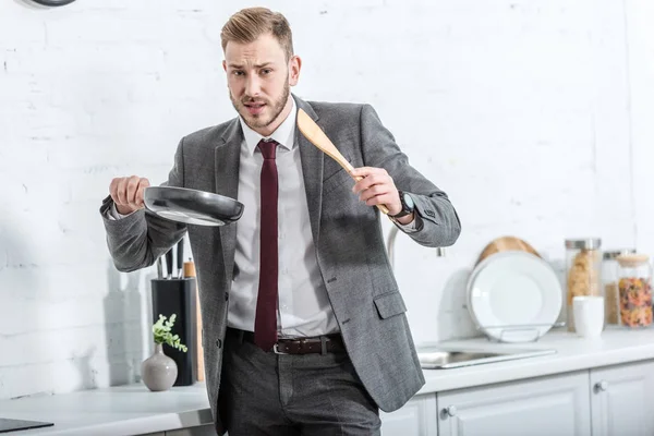Confused businessman in formal wear holding spatula with pan and getting ready to cook in kitchen — Stock Photo