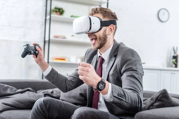 Excited businessman wearing virtual reality headset and playing video game on couch at home — Stock Photo