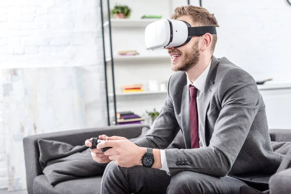 Homme d'affaires souriant portant un casque de réalité virtuelle et jouant au jeu vidéo sur le canapé à la maison — Photo de stock