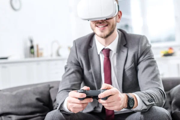 Hombre de negocios emocionado vistiendo auriculares de realidad virtual y jugando videojuego en el sofá en casa - foto de stock