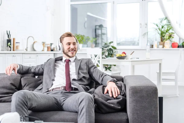 Hombre de negocios sonriente en ropa formal con auriculares y sentado en el sofá en casa - foto de stock