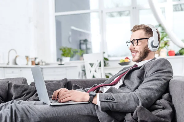 Uomo d'affari in occhiali indossando cuffie e utilizzando il computer portatile sul divano — Foto stock