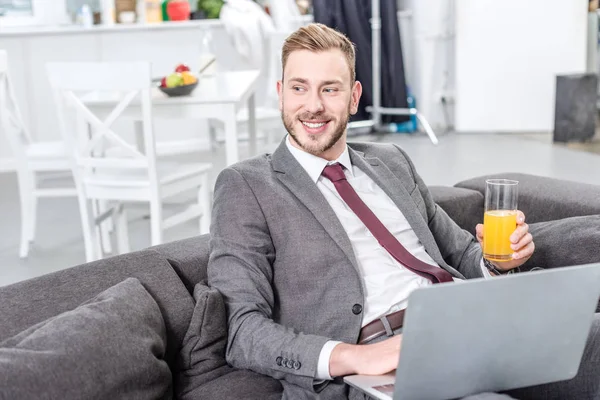 Homem de negócios sorrindo no desgaste formal beber suco de laranja e usando laptop — Fotografia de Stock