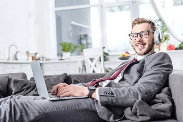 Homem de negócios sorrindo usando fones de ouvido e usando laptop no sofá — Fotografia de Stock