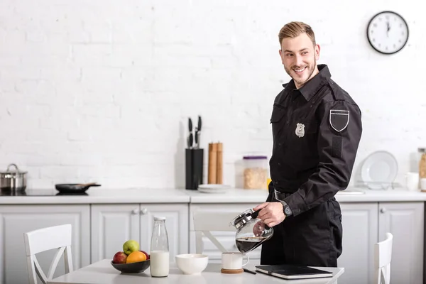 Sonriente guapo policía verter filtrado café de vidrio olla en la cocina - foto de stock