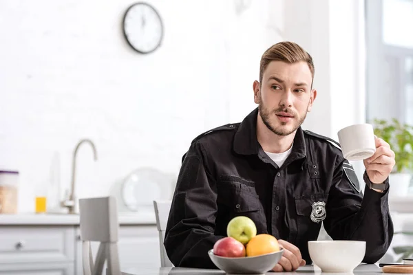 Guapo policía en la cocina sentado, sosteniendo taza de café caliente y desayunando en la mesa de la cocina - foto de stock