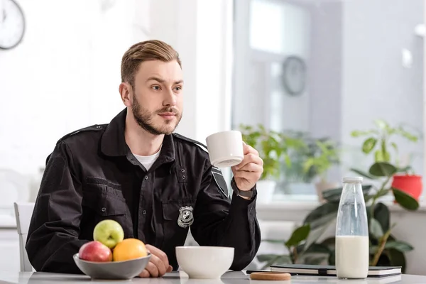 Policía en la cocina sentado, sosteniendo taza de café caliente y desayunando en la mesa de la cocina - foto de stock